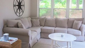 A living room with a beige sectional sofa, a white coffee table, and a large window with a view of greenery. - Close to kid-friendly Ridgevale Beach! (Chatham)