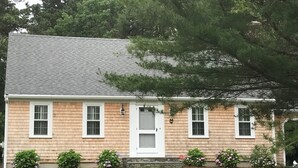 A two-story house with a gray roof, white trim, and a front door. There are three windows on the first floor and two on the second. The house is surrounded by a well-maintained lawn and several flowering shrubs. - Close to kid-friendly Ridgevale Beach! (Chatham)