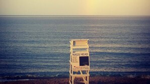 Una playa con un puesto de salvavidas identificado como 'NAGS HEAD' y un letrero que indica 'BANDERA ROJA PELIGRO'.