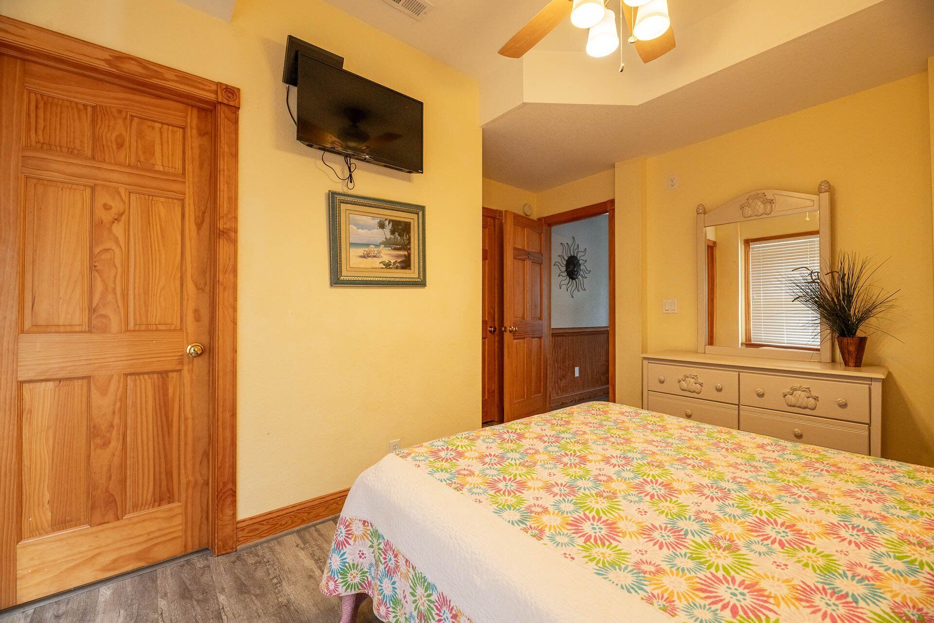 Un dormitorio con puerta de madera, televisor montado en la pared, cama con colcha estampada, cómoda con espejo y ventilador de techo.