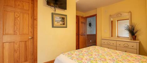 Un dormitorio con puerta de madera, televisor montado en la pared, cama con colcha estampada, cómoda con espejo y ventilador de techo.