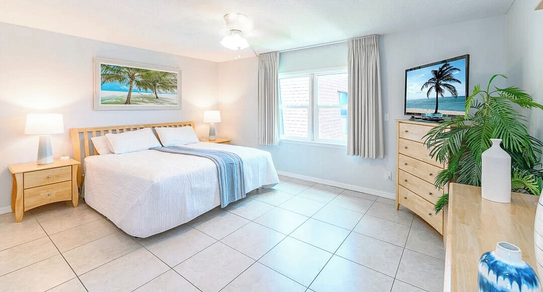 A hotel room with a bed, bedside tables, a dresser, a TV, and a window with curtains.