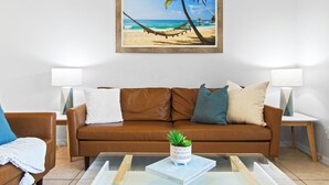 Una sala de estar con un sofá de cuero marrón, dos mesitas de noche con lámparas, una mesa de centro con un libro y una planta, y una fotografía enmarcada de una playa en la pared.
