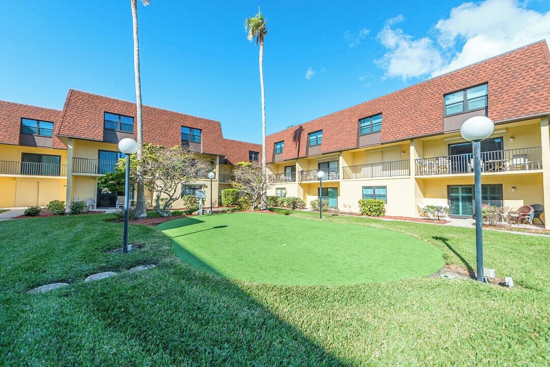 A courtyard with a green lawn, palm trees, and a building with a red roof and yellow walls.