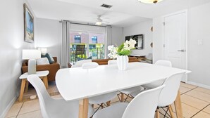 Un comedor moderno con una mesa y sillas blancas, un florero con flores y vistas al balcón.