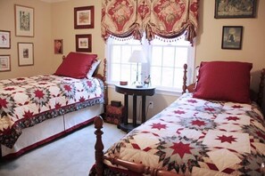 Una camera da letto con due letti, ognuno con una coperta a fiori e cuscini rossi. Si vedono quadri appesi alle pareti, una finestra con tende e un comodino con una lampada.