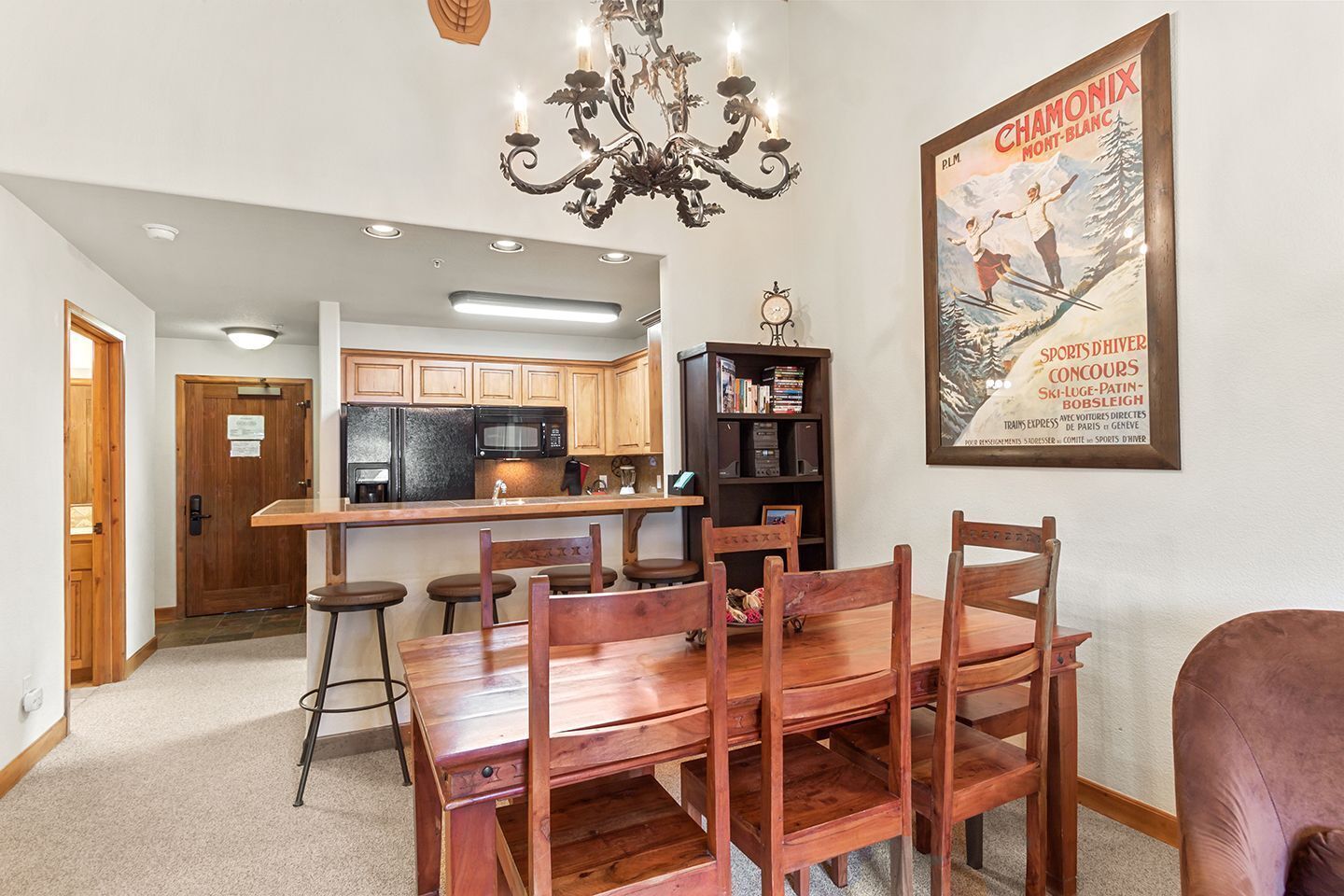 A dining area with a wooden table and chairs, a kitchen in the background, and a poster on the wall.