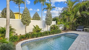 A swimming pool surrounded by tropical plants and palm trees, with a white fence and a yellow building in the background. - Cozy Cottage for 4 located steps from the beach. Heated Pool in private Patio. (Holmes Beach)