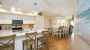 A modern kitchen with white cabinets, a large island with bar stools, and a dining area with a table and chairs. - Pool, Hot Tub, Elevator, Beach Views- Club at Mexico Beach 2D (Mexico Beach)