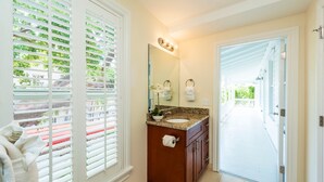 Un baño con lavabo, espejo y toallero. Hay una ventana con persianas y vistas al exterior con vegetación.