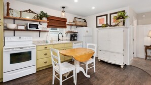 A kitchen with a wooden table and chairs, a white stove, and a white refrigerator. - Kemp & Olive Cottage: Tiny Cottage for Two Guests and Pups! (Nags Head)