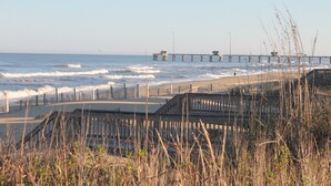 A beach with a wooden boardwalk, waves, and a pier in the background. - Kemp & Olive Cottage: Tiny Cottage for Two Guests and Pups! (Nags Head)