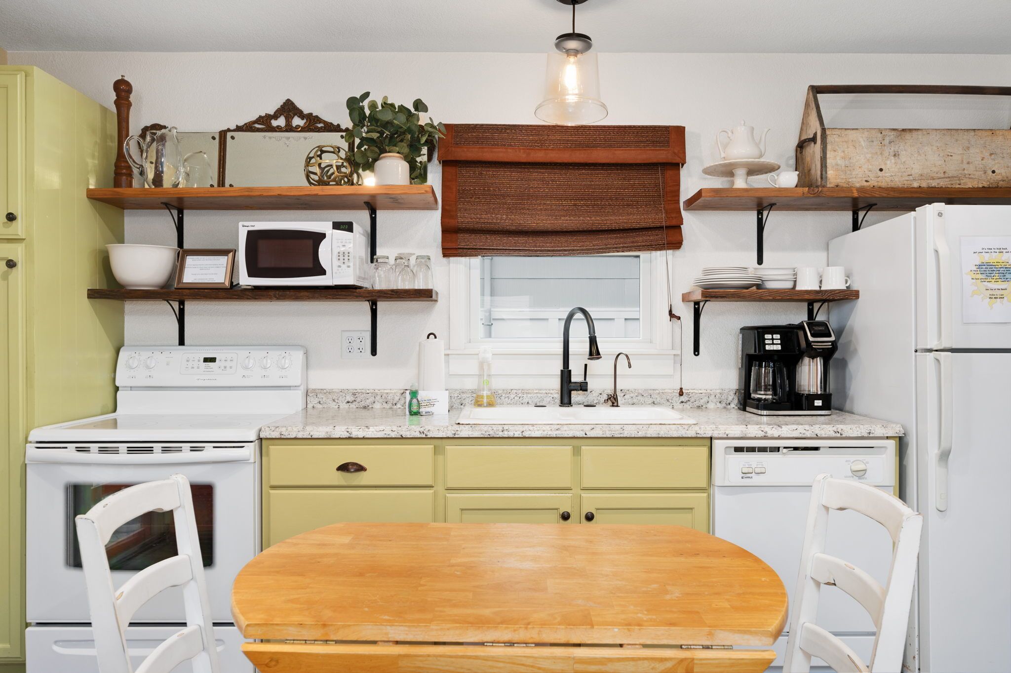 A kitchen with a wooden table, white chairs, a microwave, a stove, a sink, and shelves with various items.