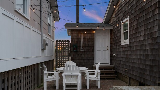 A patio with a round table and four white chairs, a wooden fence, and a house with a dark siding exterior. - Kemp & Olive Cottage: Tiny Cottage for Two Guests and Pups! (Nags Head)