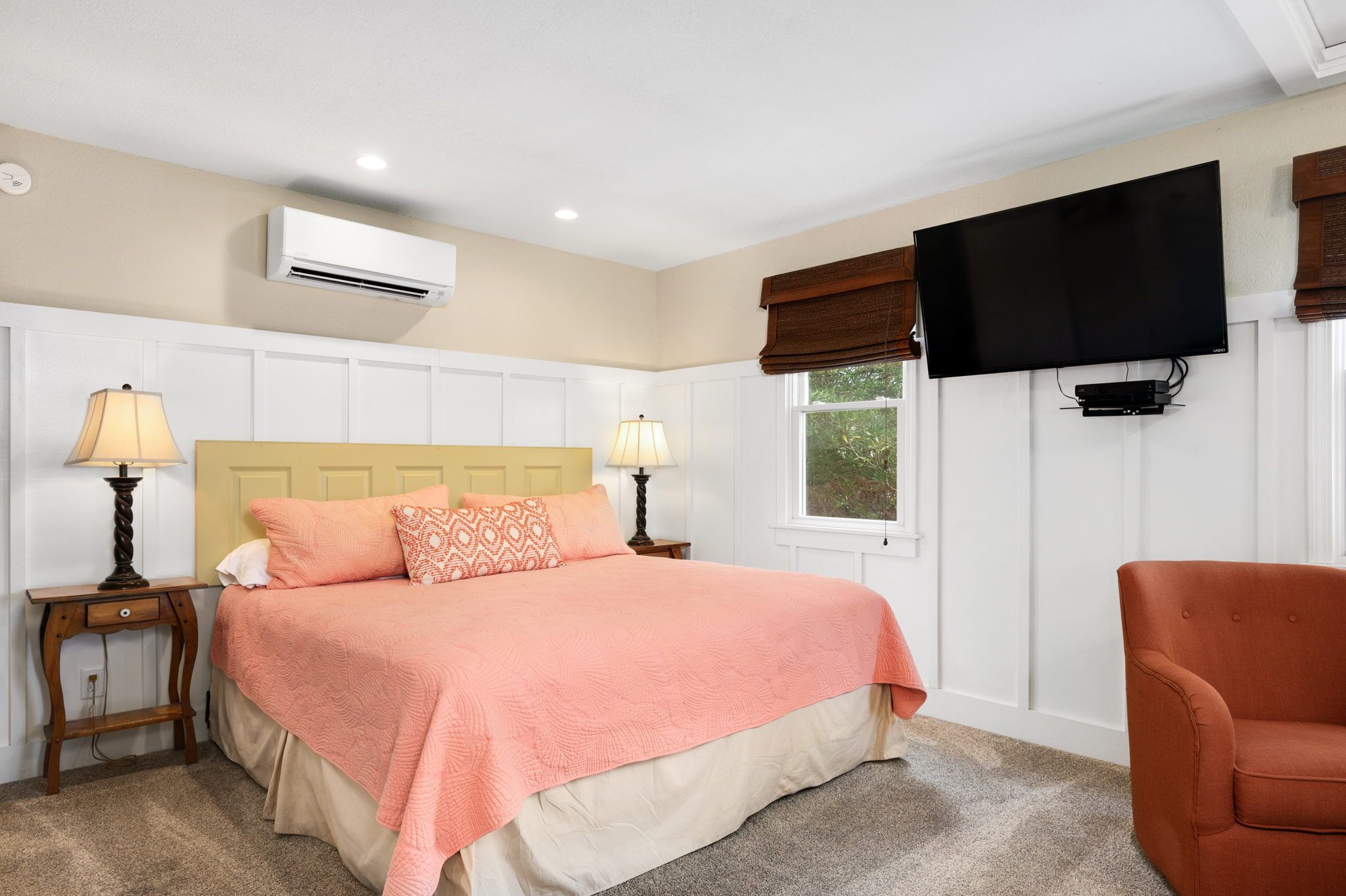 A bedroom with a bed, two bedside tables with lamps, a television mounted on the wall, and a window with blinds.