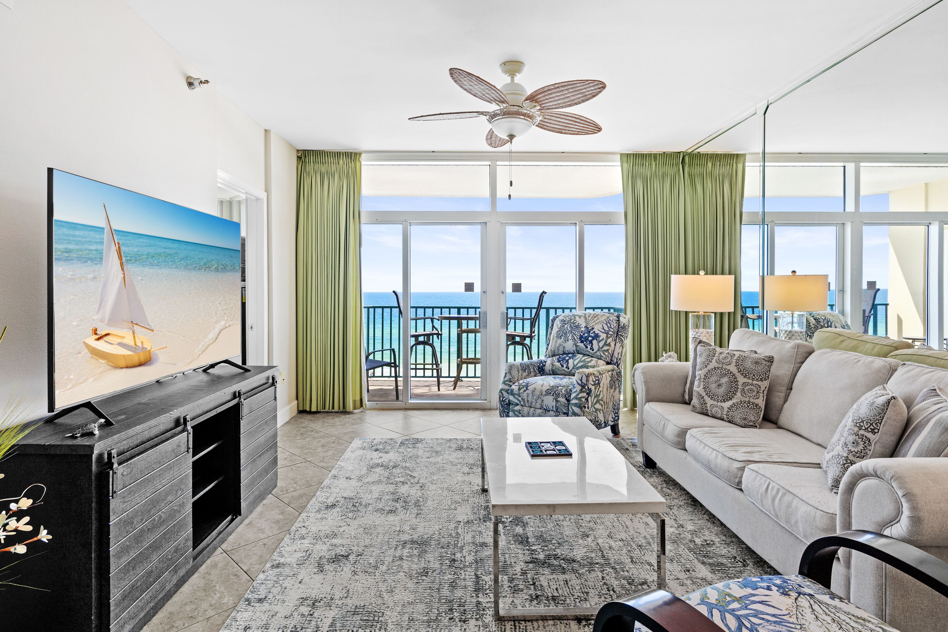 A living room with a sofa, coffee table, and a TV displaying a sailboat. There is a balcony with a view of the sea.