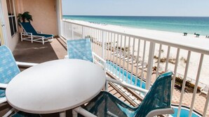 A balcony with a white table and blue chairs, overlooking a beach with a pool.