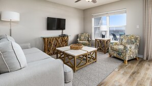 A modern living room with a ceiling fan, a flat-screen TV, a grey sofa, two patterned armchairs, and a coffee table with a woven basket.