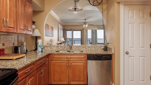 A kitchen with wooden cabinets, granite countertops, a stainless steel dishwasher, and a view of the water through large windows. - Bayfront Retreat with Pool, Perfect for 6 - PBC108 (Pensacola Beach)
