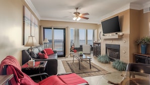 A spacious living room with a leather sofa, red cushions, a coffee table, and a fireplace. There is a large TV mounted on the wall and a ceiling fan. The room has large windows offering a view of the water. - Bayfront Retreat with Pool, Perfect for 6 - PBC108 (Pensacola Beach)