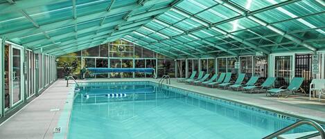 A swimming pool with a glass ceiling, lounge chairs, and a view of a building outside.