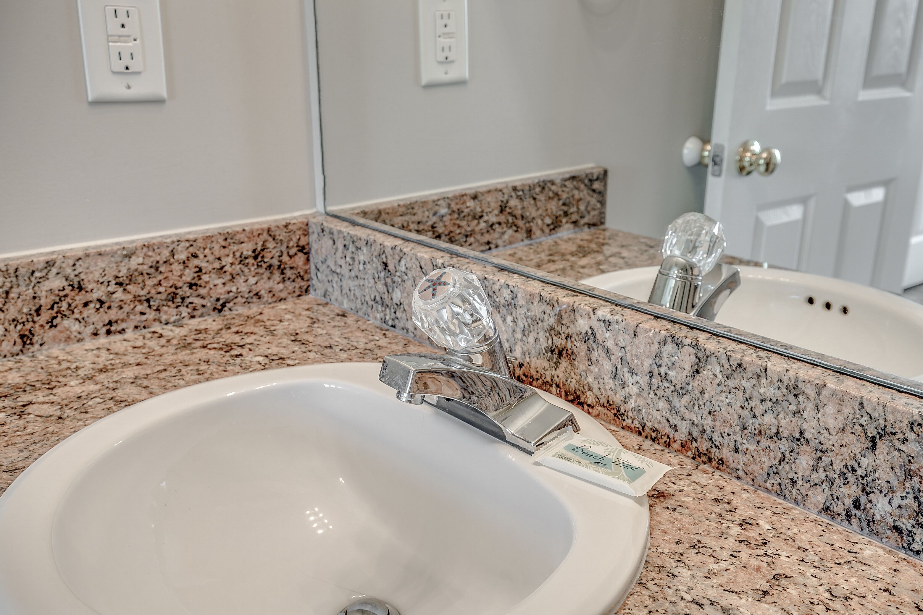 A bathroom with a white sink, granite countertop, and a mirror reflecting the sink and door.