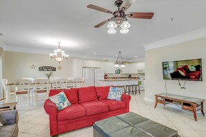 Ein geräumiges Wohnzimmer mit einem roten Sofa, einem Glastisch, einem Ledersitzhocker, einem Deckenventilator und einem an der Wand befestigten Fernseher.
