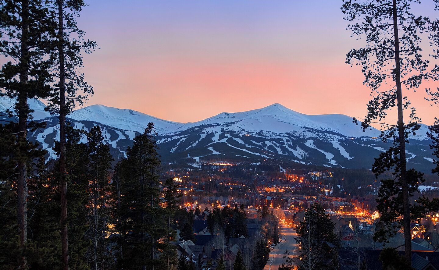 A town nestled between snow-covered mountains at dusk with visible buildings and roads.