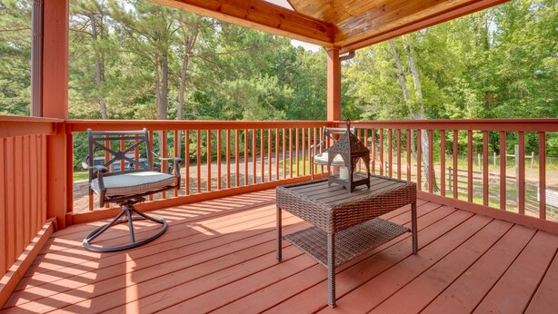 Una terraza techada al aire libre con una mesa y una silla de madera, rodeada de barandas de madera y con vista a un área boscosa.