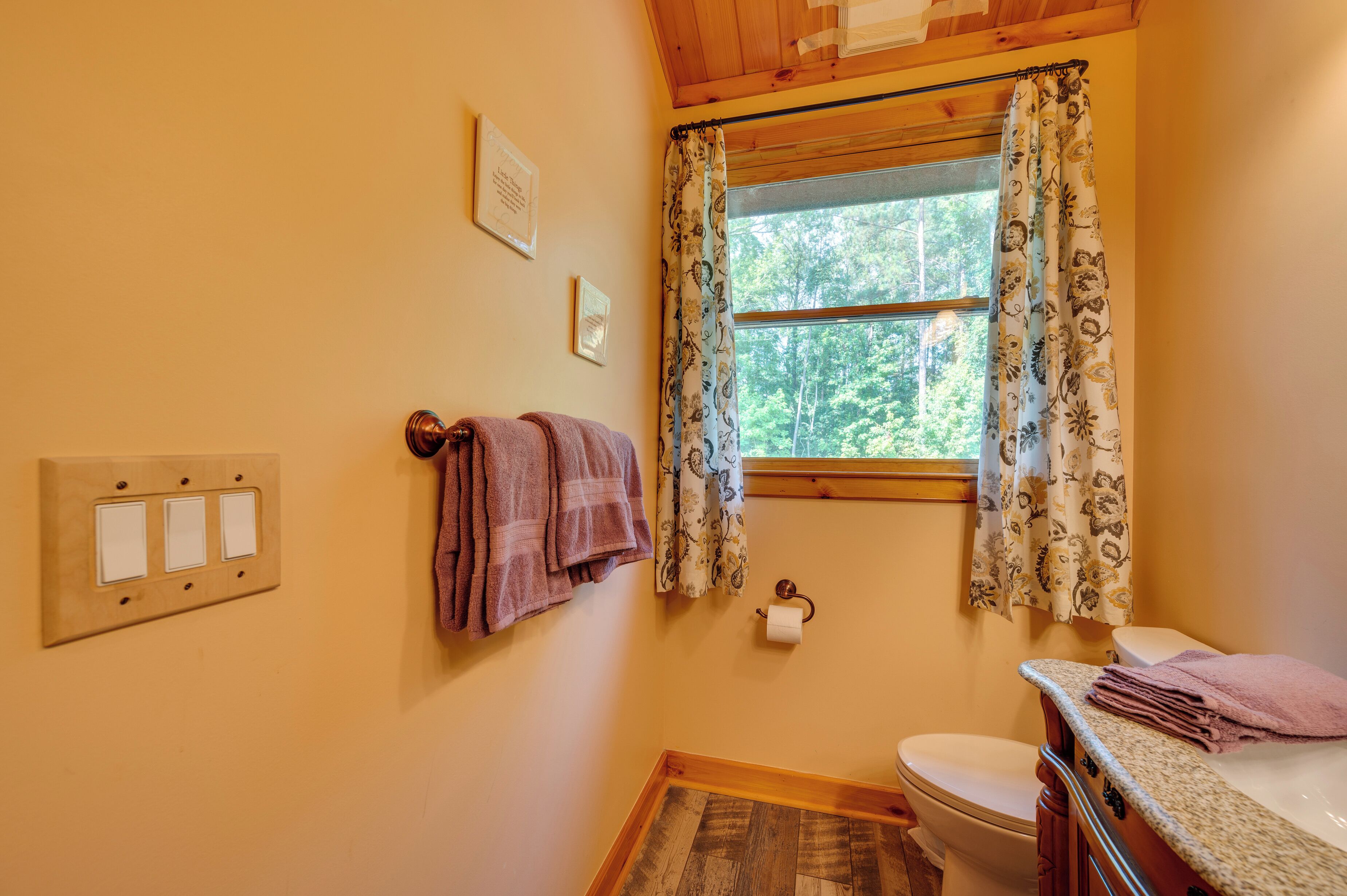 Une salle de bain avec un plafond en bois, une fenêtre ornée de rideaux à motifs floraux, des toilettes et un lavabo avec un porte-serviettes.