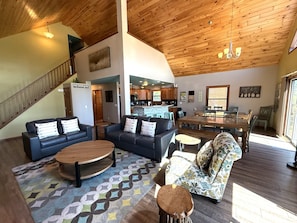 A spacious living area with a wooden ceiling, a dining table, sofas, and a kitchen in the background.