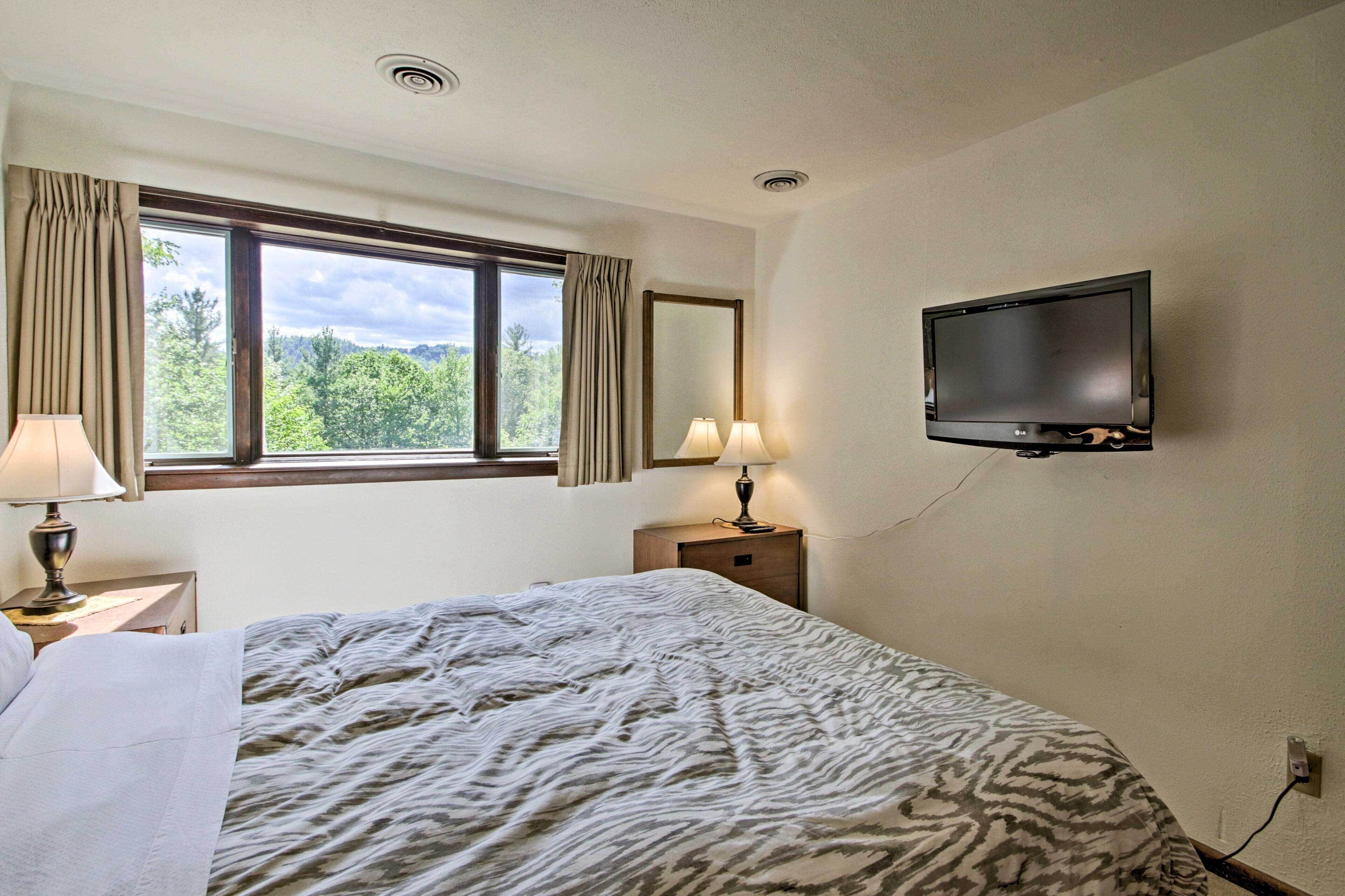 A hotel room with a bed, a TV mounted on the wall, a window with curtains, and a nightstand with a lamp.