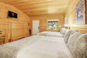 A wooden cabin room with two beds, a television, a window with a tree view, and a door.
