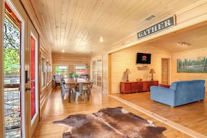 A spacious room with a wooden ceiling, a dining area with chairs and a table, a blue sofa, a mounted television, and a sign that reads 'GATHERER'.