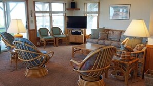 A cozy living room with wicker furniture, a flat-screen TV, and a large window providing natural light. - KD906- Semi-OF, 4 BRs, 2 Living Areas, Crow's Nest and Beautiful Views! (Kill Devil Hills)