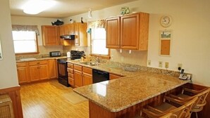 A kitchen with wooden cabinets, a granite countertop, a dishwasher, and a dining area with chairs. - KD906- Semi-OF, 4 BRs, 2 Living Areas, Crow's Nest and Beautiful Views! (Kill Devil Hills)