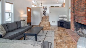 Una sala de estar moderna con un sofá gris, una mesa de centro y un televisor sobre un mueble bajo. Se ve una escalera que conduce a un nivel superior y una chimenea de ladrillo a la derecha.