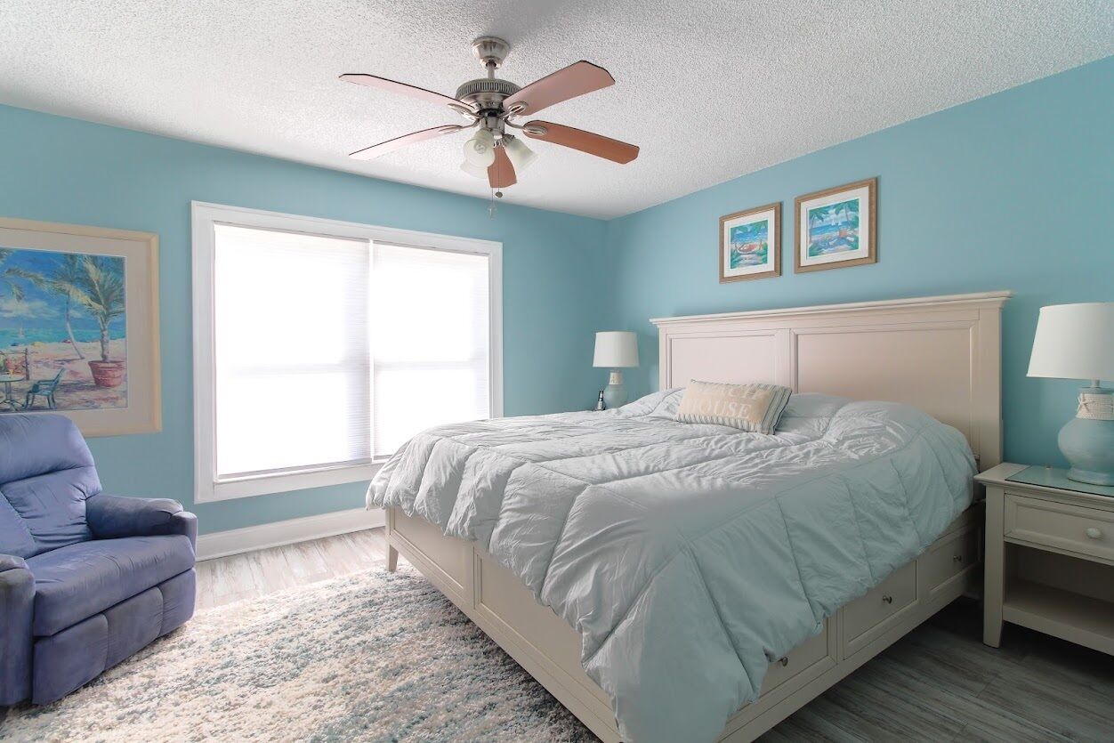 Un dormitorio con cama, ventilador de techo, ventana, silla y mesita de noche.