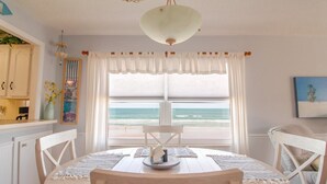 Un comedor con vista al océano, que incluye una mesa redonda, sillas blancas, una lámpara colgante y una ventana con cortinas blancas.