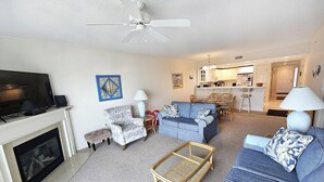 Una sala de estar con chimenea, televisor, ventilador de techo, sofá, mesa de centro y un comedor al fondo.
