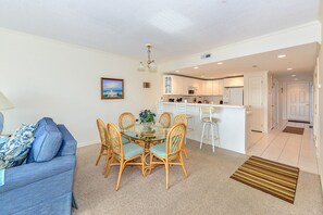 Cozy living space with a dining area featuring a glass-top table and four chairs, a kitchenette with a refrigerator, and a framed picture on the wall.