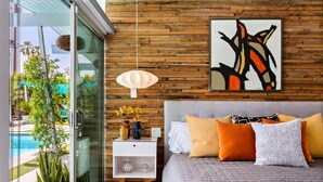 A modern bedroom with a glass door leading to a pool, a wooden wall, and a bed with multiple pillows.
