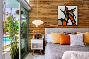 A modern bedroom with a glass door leading to a pool, a wooden wall, and a bed with multiple pillows.