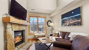 Una sala de estar con chimenea de piedra, un ventanal con vistas a montañas nevadas, un sofá de cuero marrón y un televisor montado.