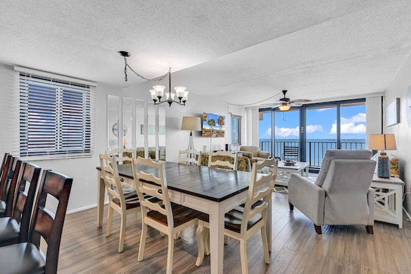 A spacious living room with a large wooden dining table surrounded by chairs, a sofa, and a balcony with a view of the ocean.