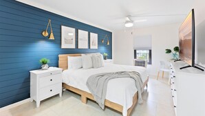 A bedroom with a bed, bedside tables, a TV, and a window with blinds. - ☀️ Sun-Kissed Escape  l  Oceanfront Condo w/ Private Balcony l Pool, Hot Tub & Tennis (Cape Canaveral)