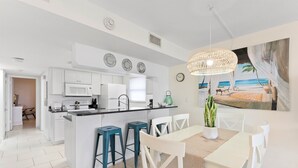 A modern kitchen with white cabinets, a central island with stools, and a dining area with a table and chairs. - ☀️ Sun-Kissed Escape  l  Oceanfront Condo w/ Private Balcony l Pool, Hot Tub & Tennis (Cape Canaveral)