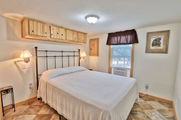 Un dormitorio con cama, lámparas de noche, una ventana con vistas, un armario de madera y un cuadro en la pared.