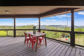 A covered outdoor area with a wooden deck, a table with red chairs, and a view of a hilly landscape. - On Top of the World  I Minutes from beautiful Canyon Lake (Wimberley)