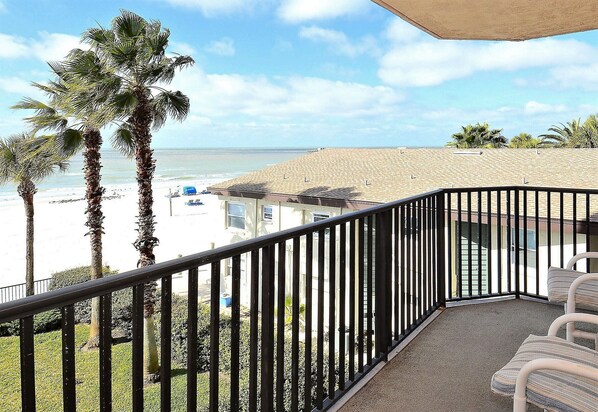 A balcony with a view of the beach, palm trees, and a building in the background.